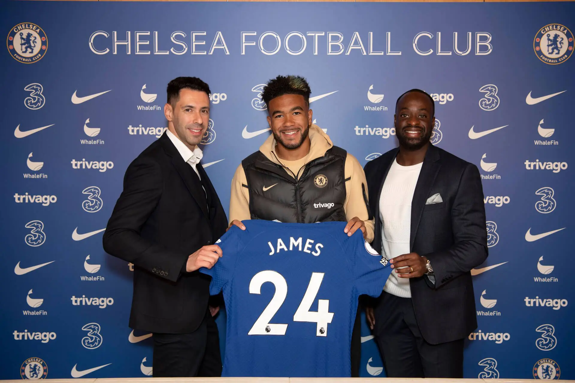 Mike-+-Reece-James-new (1) Three men holding a soccer jersey in front of a blue wall.